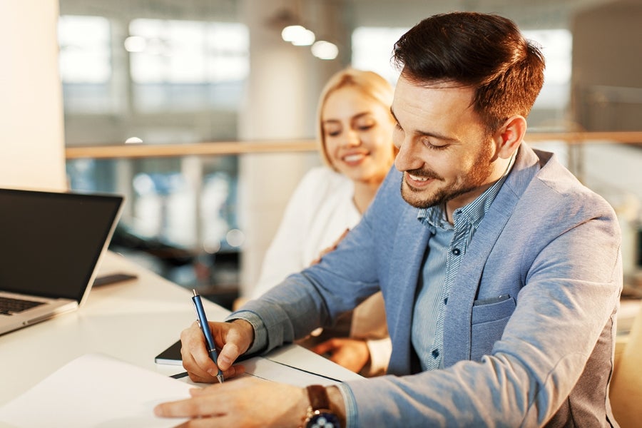 Person seated at a desk signing a document with a pen while another person sits nearby in the background
