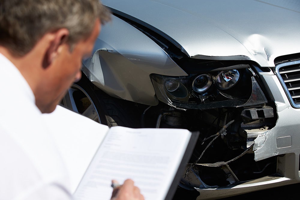 An insurance adjuster reviews damage to the front a silver car, with a crumpled hood, fender, and broken headlight