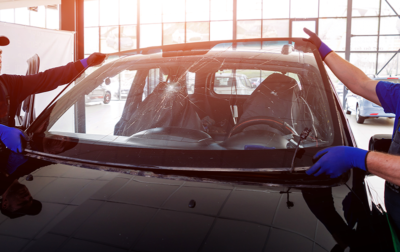 Two technicians wearing blue gloves are removing a severely cracked windshield from a black car in a garage