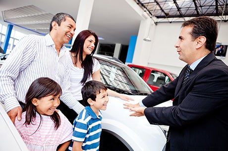 A car salesman speaks animatedly to a laughing family of four (parents, daughter, son) at a dealership
