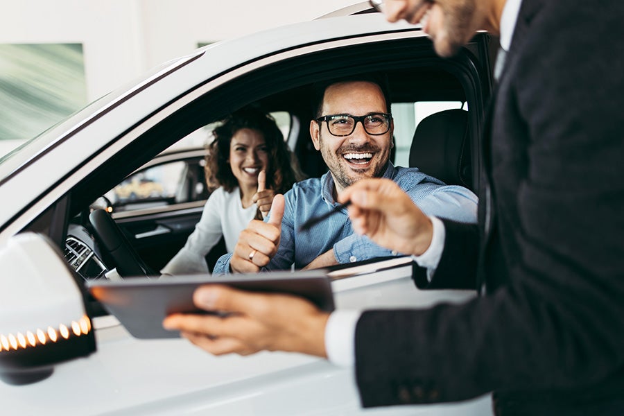 Smiling couple in a white car giving thumbs up to a salesperson holding a tablet