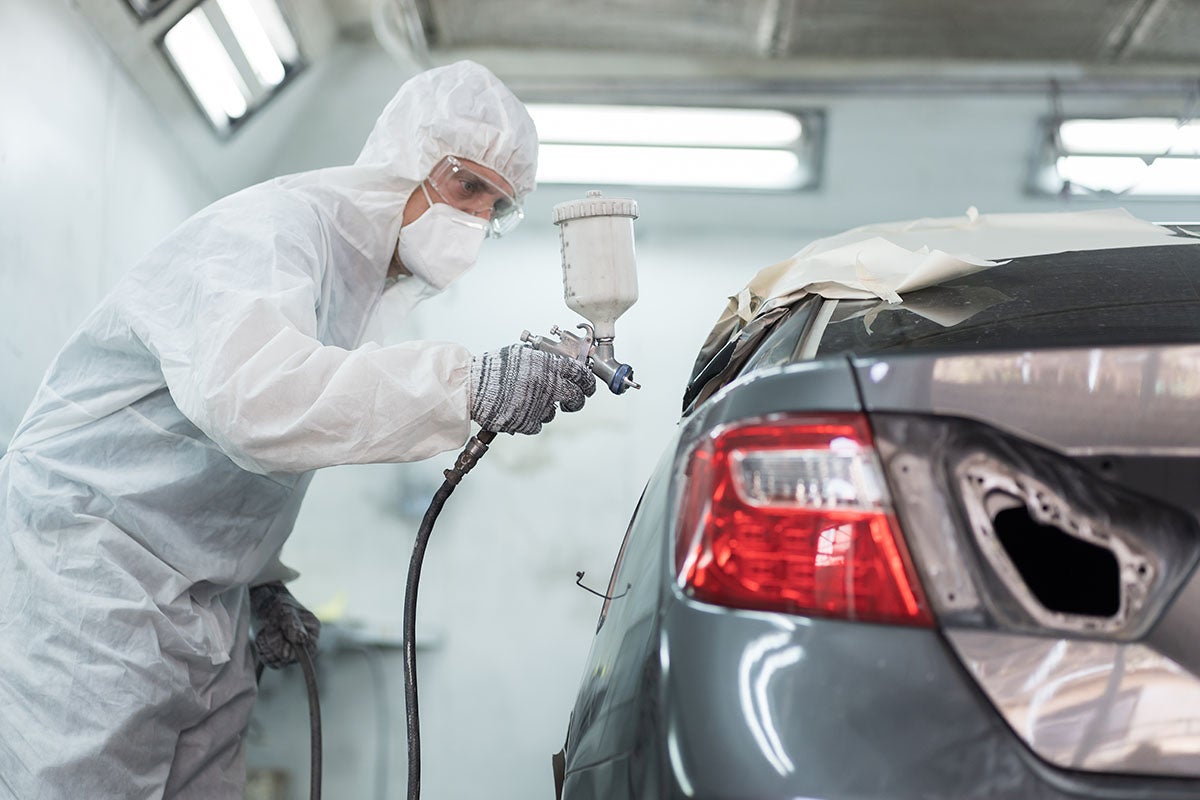 A person in a white protective suit and mask spray paints the rear of a grey car in a garage