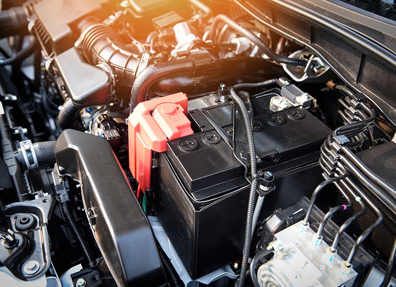 Close-up view of a vehicle engine compartment featuring a black automotive battery with a red positive terminal cover in the foreground